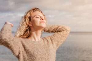 Woman in brown sweater by the ocean closing her eyes facing the sun